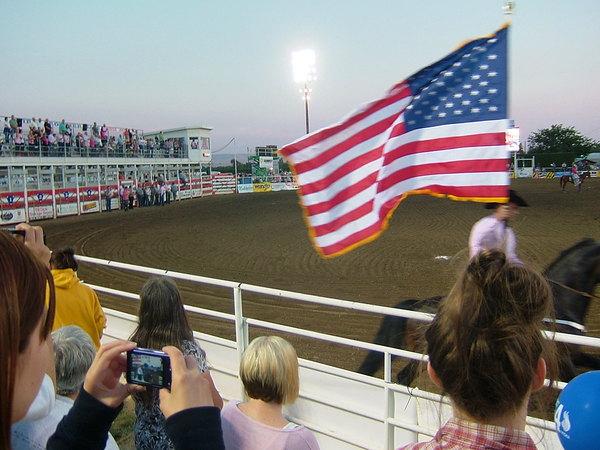 男性がアメリカの国旗を手に持ち、馬に乗っているところを観客が見ている写真